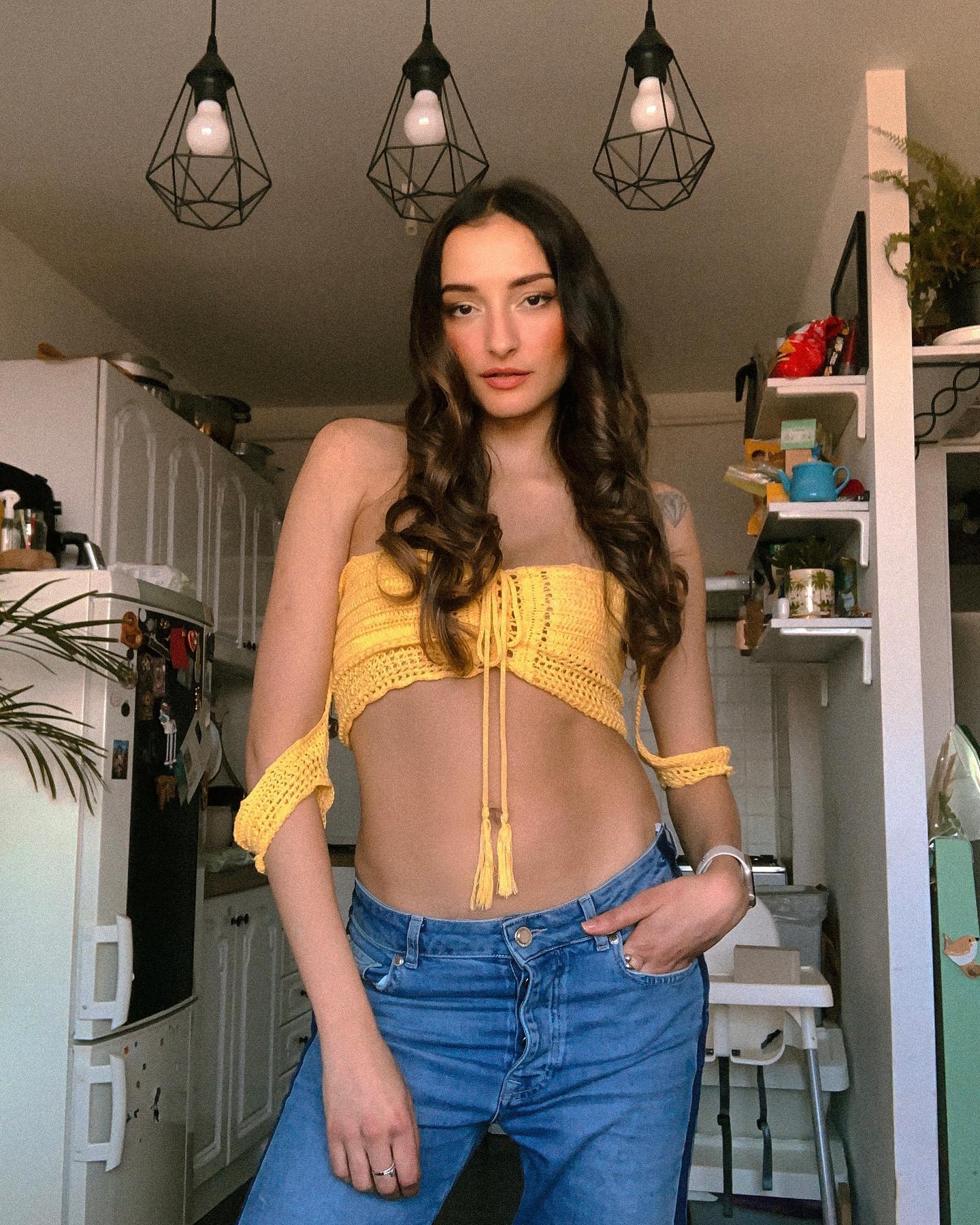 Rafaela in a yellow crochet top and high-waist jeans, sunlit kitchen with three geometric pendant lights overhead and a leafy plant.