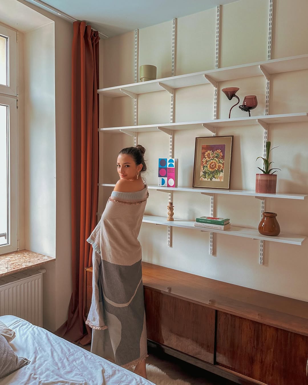 Rafaela wrapped in a geometric-pattern throw, sunlit room with white shelving, framed sunflower painting, orange linen curtain — slow lifestyle interior, November 2023.